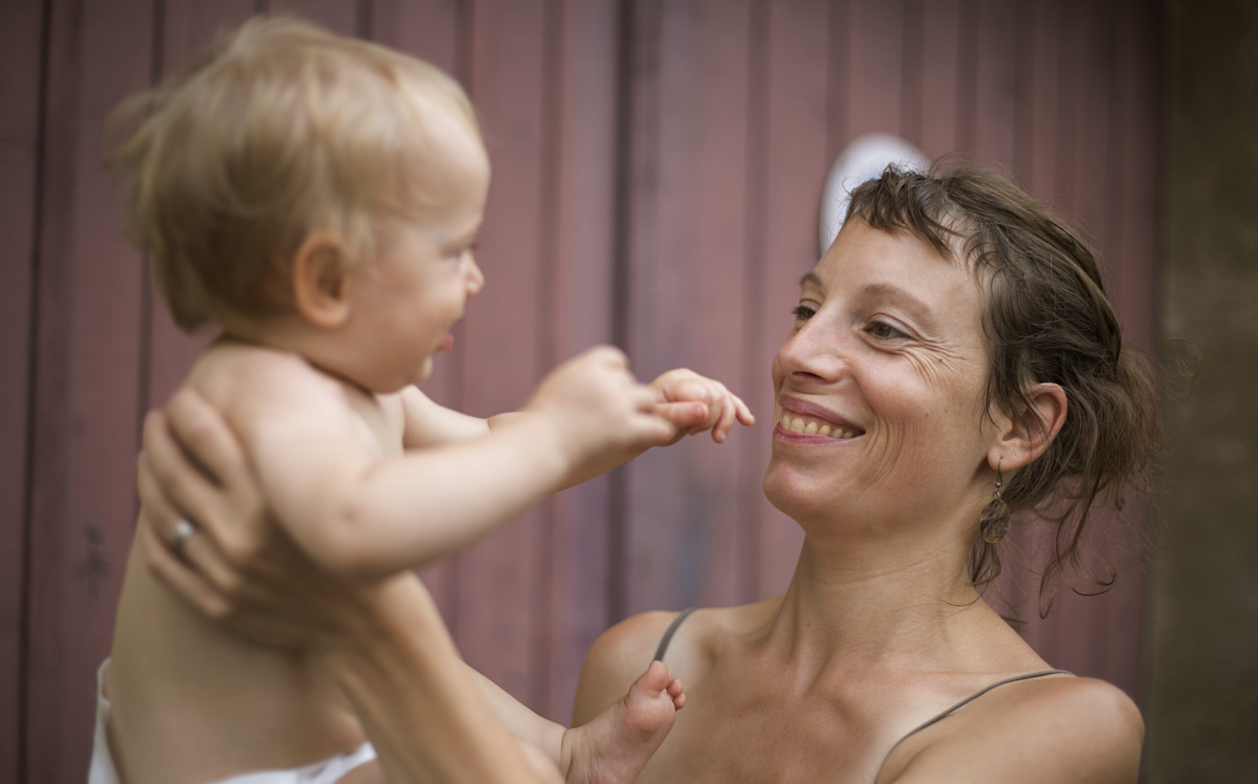 Une mère tient son bébé face à elle, tous deux souriants, devant une palissade de bois rose