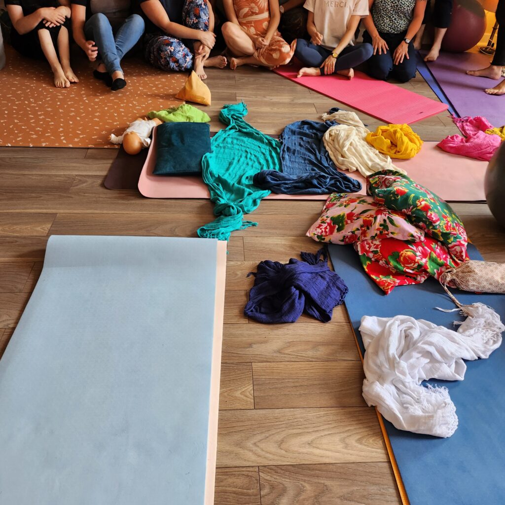Vue de dessus d’une salle de transmission : tapis disposés en cercle, tissus rebozo pliés par couleur, coussins fleuris et poupon d’exercice ; les participantes sont assises en périphérie