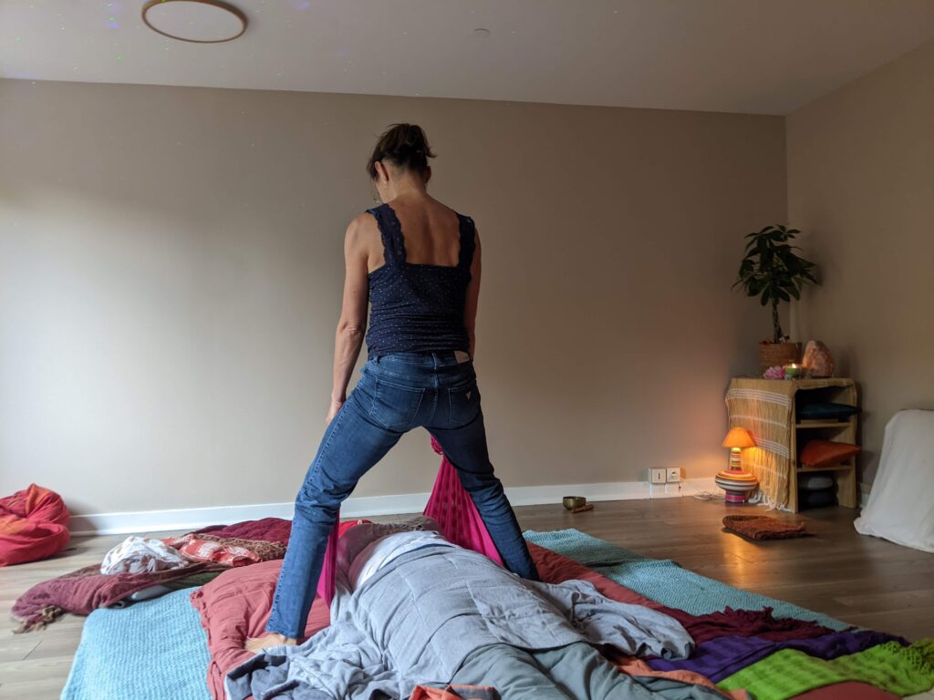 La praticienne, debout et vue de dos, tend un tissu rose au-dessus d’une personne allongée et couverte, dans une pièce éclairée par des lampes